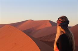 Pale blue sky, huge red san dunes. In the foreground a Young woman with Long blond hair and a colourful headband Looks the the horizon. 